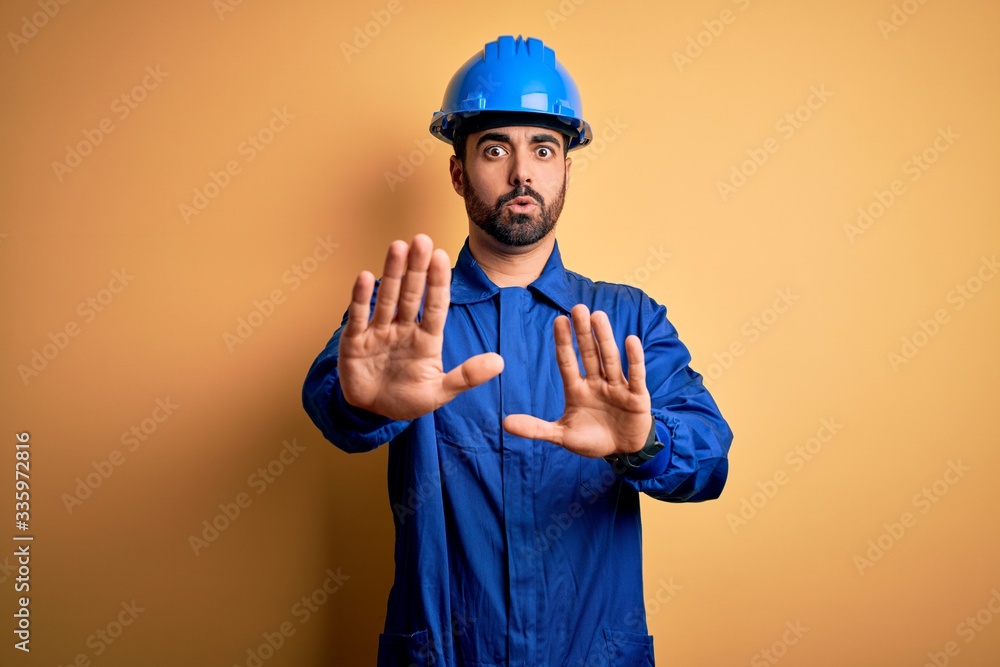 Mechanic man with beard wearing blue uniform and safety helmet over yellow background Moving away hands palms showing refusal and denial with afraid and disgusting expression. Stop and forbidden.