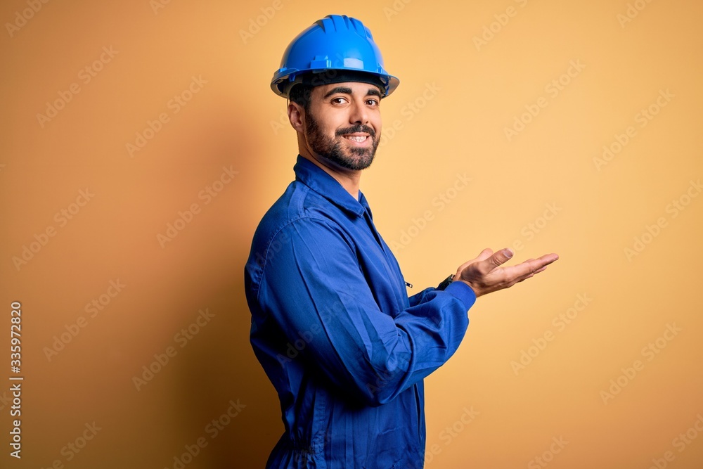 Mechanic man with beard wearing blue uniform and safety helmet over yellow background pointing aside with hands open palms showing copy space, presenting advertisement smiling excited happy