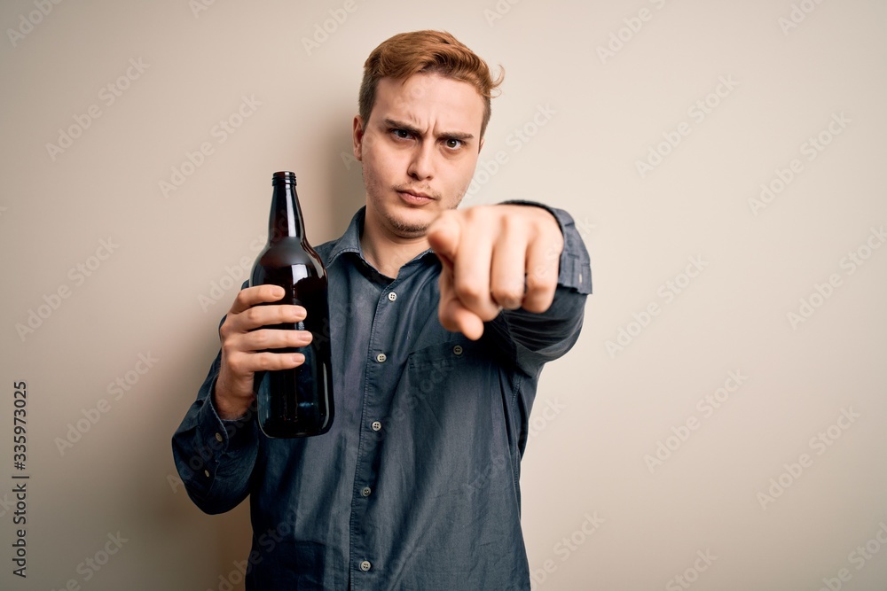 Young handsome redhead man drinking bottle of beer over isolated white background pointing with finger to the camera and to you, confident gesture looking serious Young handsome redhead man drinking bottle of beer over isolated white background pointing with finger to the camera and to you, confident gesture looking serious