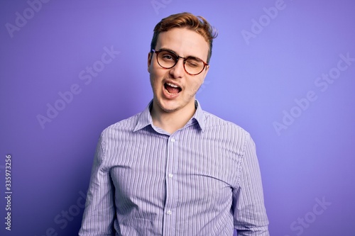 Young handsome redhead man wearing casual shirt and glasses over purple background winking looking at the camera with sexy expression, cheerful and happy face.
