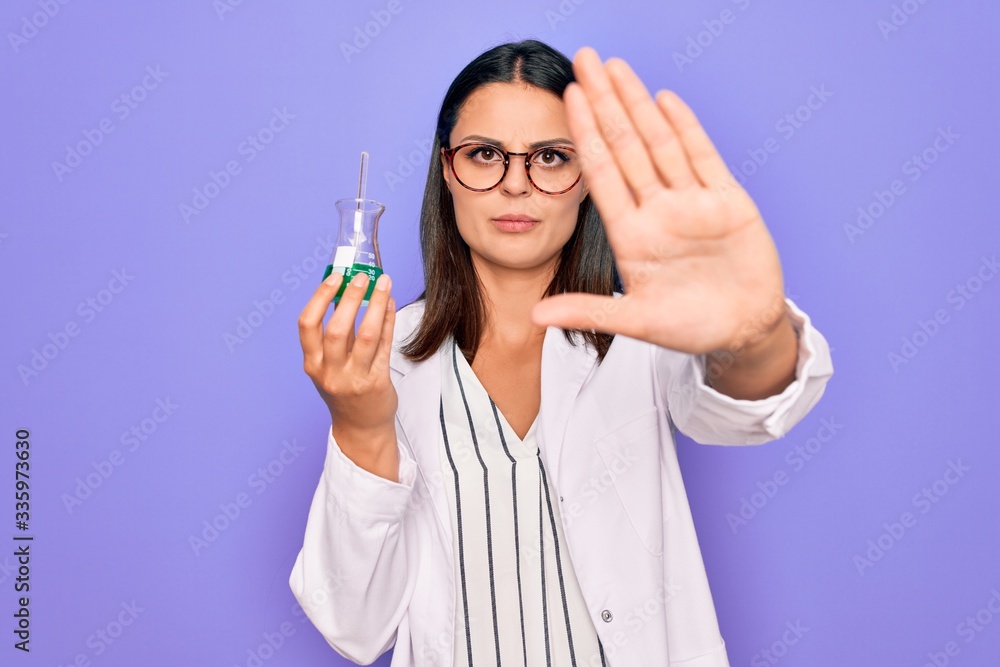 Young beautiful brunette scientist woman wearing coat and glasses holding test tube with open hand doing stop sign with serious and confident expression, defense gesture Young beautiful brunette scientist woman wearing coat and glasses holding test tube with open hand doing stop sign with serious and confident expression, defense gesture