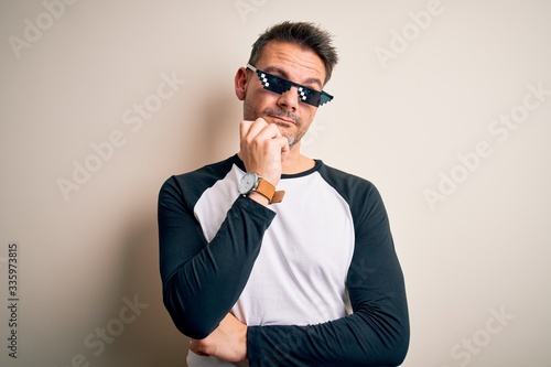 Young handsome man wearing funny thug life sunglasses meme over white background with hand on chin thinking about question, pensive expression. Smiling with thoughtful face. Doubt concept.
