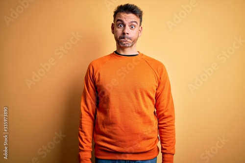 Young handsome man wearing orange casual sweater standing over isolated yellow background puffing cheeks with funny face. Mouth inflated with air, crazy expression.