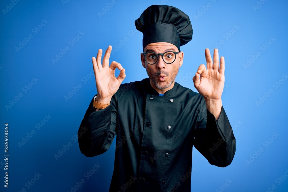 Young handsome chef man wearing cooker uniform and hat over isolated blue background looking surprised and shocked doing ok approval symbol with fingers. Crazy expression Young handsome chef man wearing cooker uniform and hat over isolated blue background looking surprised and shocked doing ok approval symbol with fingers. Crazy expression