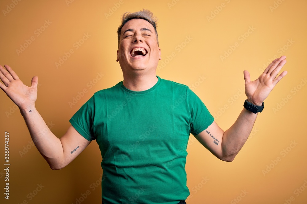 Fototapeta premium Young handsome modern man wearing casual green t-shirt over yellow background celebrating mad and crazy for success with arms raised and closed eyes screaming excited. Winner concept