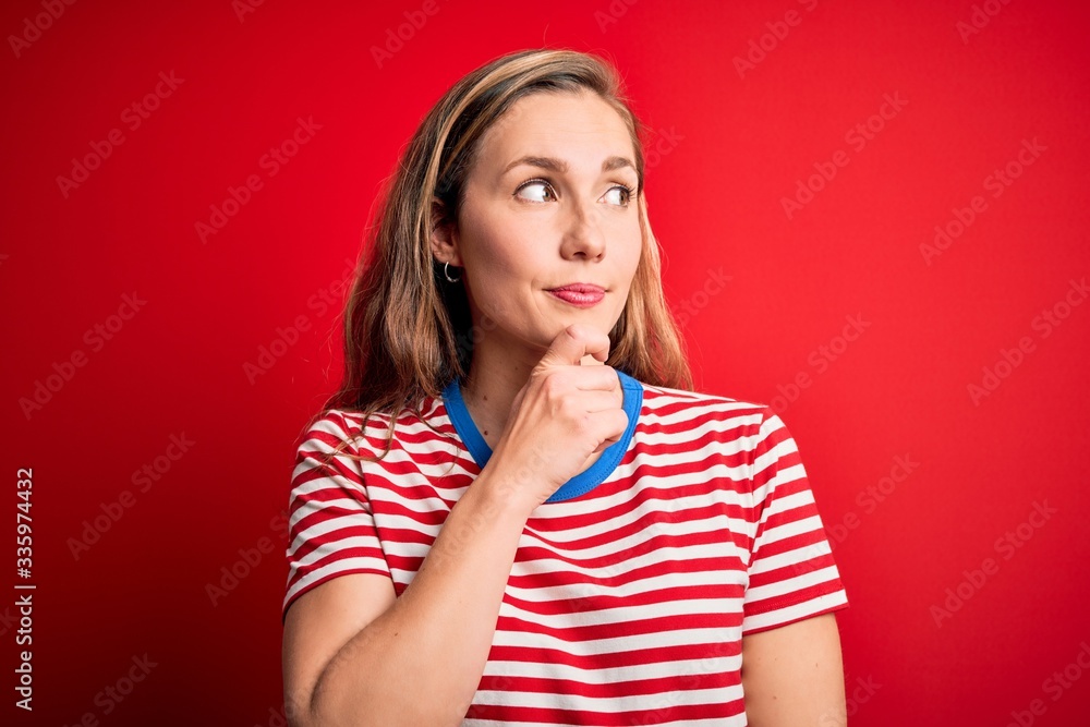 Young beautiful blonde woman wearing casual striped t-shirt over isolated red background with hand on chin thinking about question, pensive expression. Smiling with thoughtful face. Doubt concept. Young beautiful blonde woman wearing casual striped t-shirt over isolated red background with hand on chin thinking about question, pensive expression. Smiling with thoughtful face. Doubt concept.