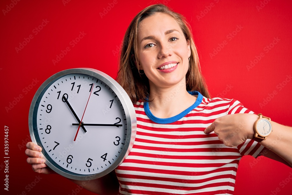 Young beautiful blonde woman holding big clock standing over isolated red background with surprise face pointing finger to himself Young beautiful blonde woman holding big clock standing over isolated red background with surprise face pointing finger to himself