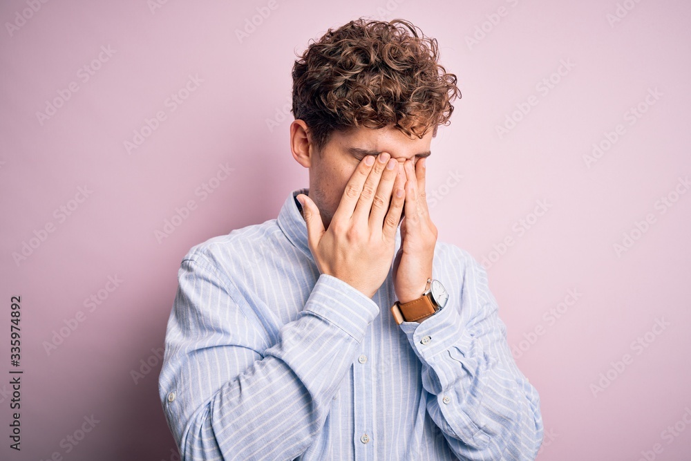Young blond handsome man with curly hair wearing striped shirt over white background rubbing eyes for fatigue and headache, sleepy and tired expression. Vision problem Young blond handsome man with curly hair wearing striped shirt over white background rubbing eyes for fatigue and headache, sleepy and tired expression. Vision problem