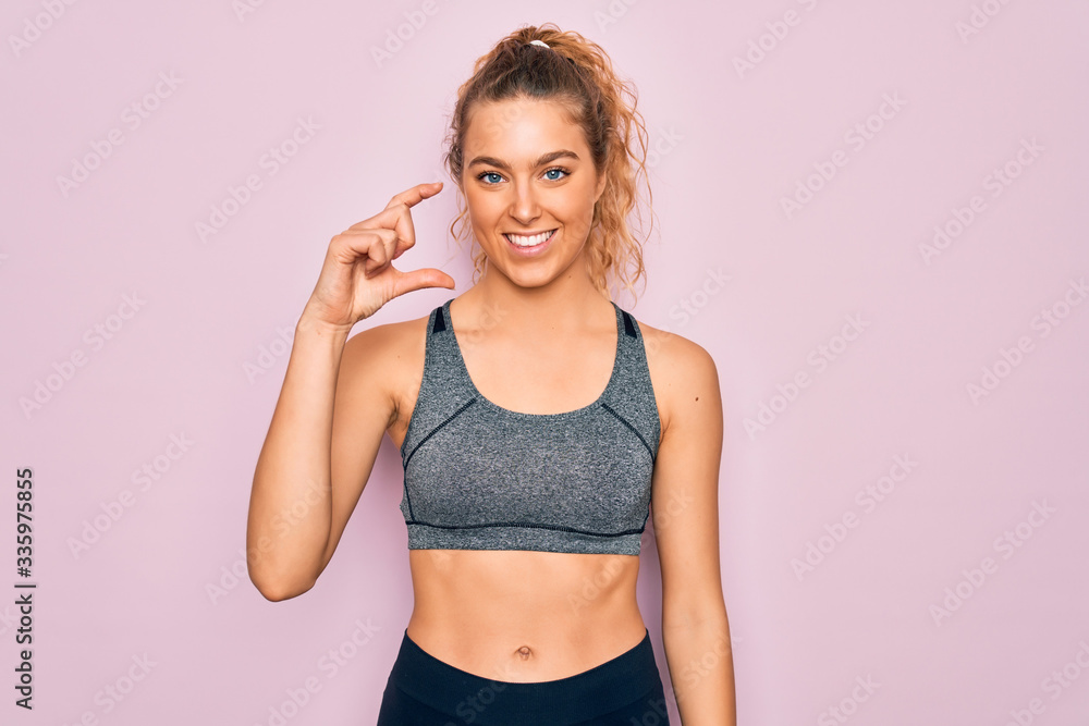 Young beautiful blonde sportswoman with blue eyes doing exercise wearing sportswear smiling and confident gesturing with hand doing small size sign with fingers looking and the camera. Measure Young beautiful blonde sportswoman with blue eyes doing exercise wearing sportswear smiling and confident gesturing with hand doing small size sign with fingers looking and the camera. Measure