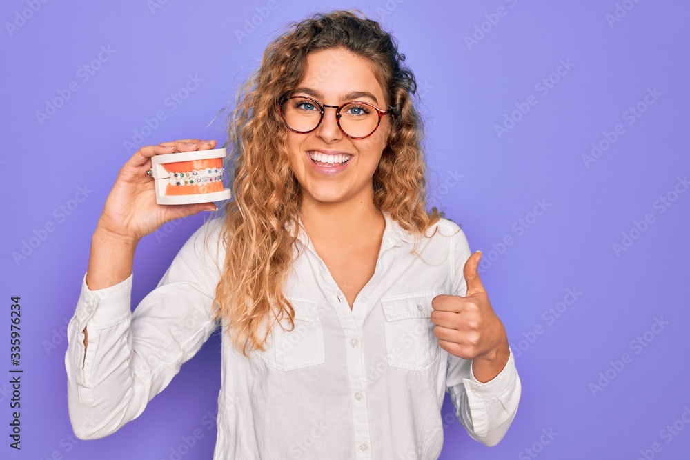 Beautiful blonde woman with blue eyes holding plastic dentrure teeth over purple background happy with big smile doing ok sign, thumb up with fingers, excellent sign Beautiful blonde woman with blue eyes holding plastic dentrure teeth over purple background happy with big smile doing ok sign, thumb up with fingers, excellent sign