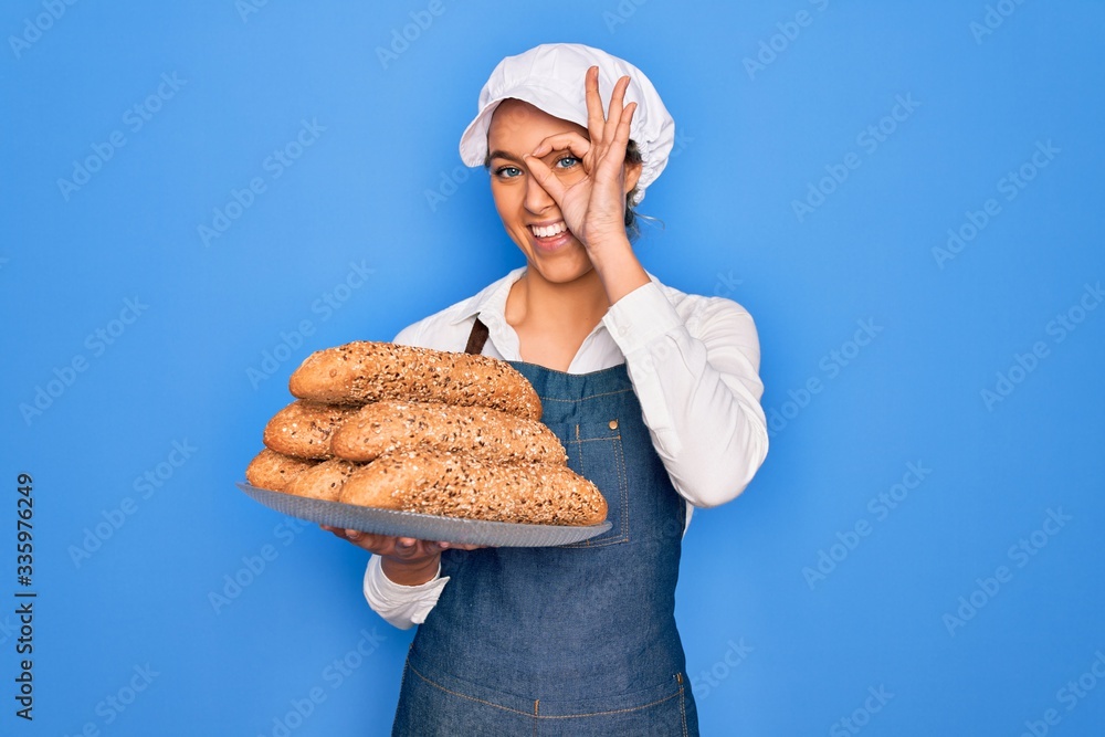 Young beautiful blonde baker woman with blue eyes holding tray with wholemeal bread cereal with happy face smiling doing ok sign with hand on eye looking through fingers Young beautiful blonde baker woman with blue eyes holding tray with wholemeal bread cereal with happy face smiling doing ok sign with hand on eye looking through fingers