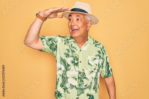 Middle age senior grey-haired man wearing summer hat and floral shirt on beach vacation very happy and smiling looking far away with hand over head. Searching concept.