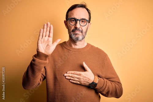 Middle age hoary man wearing brown sweater and glasses over isolated yellow background Swearing with hand on chest and open palm, making a loyalty promise oath photo