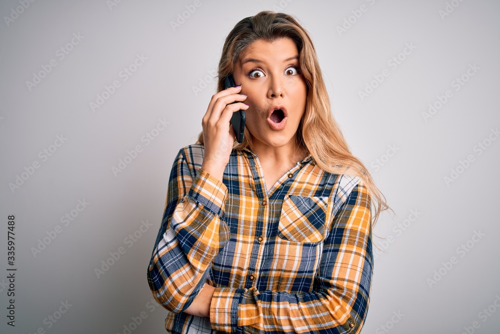 Beautiful blonde woman having conversation talking on the smartphone over white background scared in shock with a surprise face, afraid and excited with fear expression Beautiful blonde woman having conversation talking on the smartphone over white background scared in shock with a surprise face, afraid and excited with fear expression