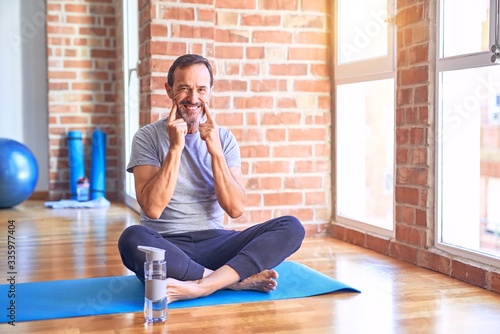 Middle age handsome sportman sitting on mat doing stretching yoga exercise at gym Smiling with open mouth  fingers pointing and forcing cheerful smile
