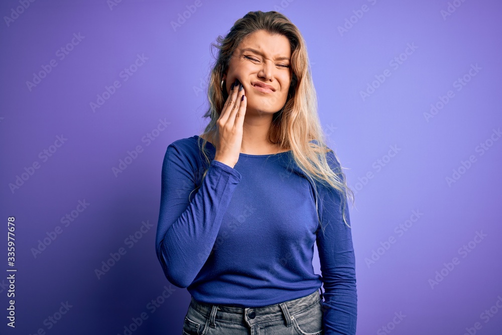 Young beautiful blonde woman wearing casual t-shirt over isolated purple background touching mouth with hand with painful expression because of toothache or dental illness on teeth. Dentist Young beautiful blonde woman wearing casual t-shirt over isolated purple background touching mouth with hand with painful expression because of toothache or dental illness on teeth. Dentist