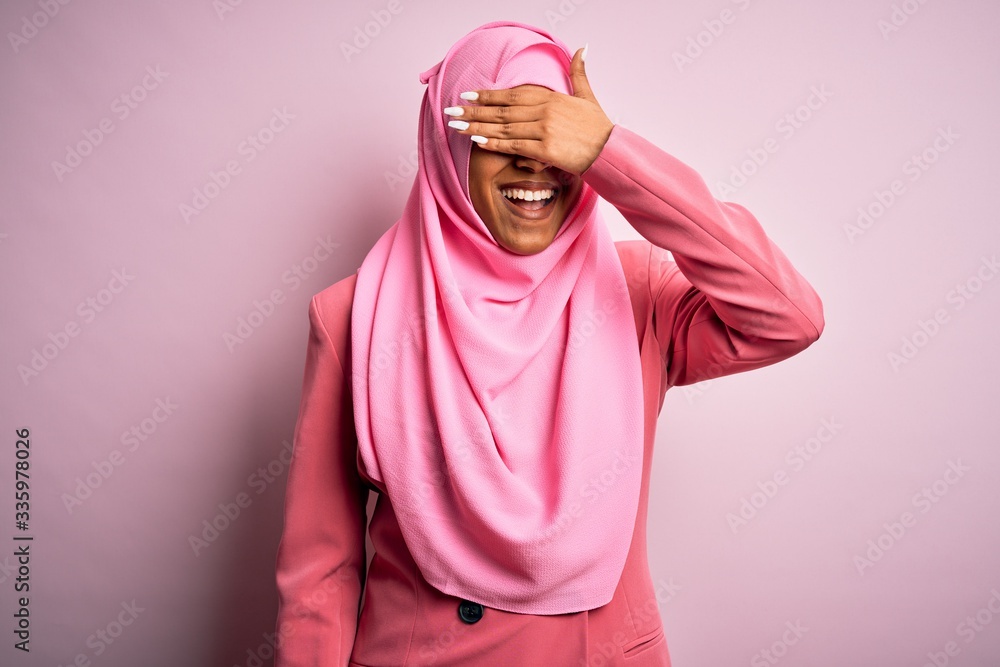 Young African American afro woman wearing muslim hijab over isolated pink background smiling and laughing with hand on face covering eyes for surprise. Blind concept. Young African American afro woman wearing muslim hijab over isolated pink background smiling and laughing with hand on face covering eyes for surprise. Blind concept.