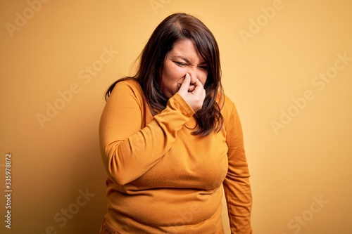 Beautiful brunette plus size woman wearing casual sweater over isolated yellow background smelling something stinky and disgusting, intolerable smell, holding breath with fingers on nose. Bad smell © Krakenimages.com