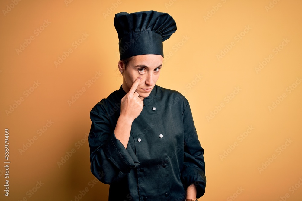 Young beautiful chef woman wearing cooker uniform and hat standing over yellow background Pointing to the eye watching you gesture, suspicious expression Young beautiful chef woman wearing cooker uniform and hat standing over yellow background Pointing to the eye watching you gesture, suspicious expression