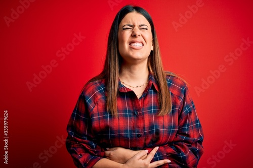 Young beautiful woman wearing casual shirt over red background with hand on stomach because indigestion, painful illness feeling unwell. Ache concept. © Krakenimages.com