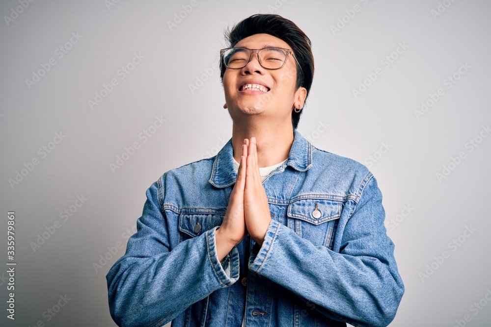 Young handsome chinese man wearing denim jacket and glasses over white background begging and praying with hands together with hope expression on face very emotional and worried. Begging. Young handsome chinese man wearing denim jacket and glasses over white background begging and praying with hands together with hope expression on face very emotional and worried. Begging.