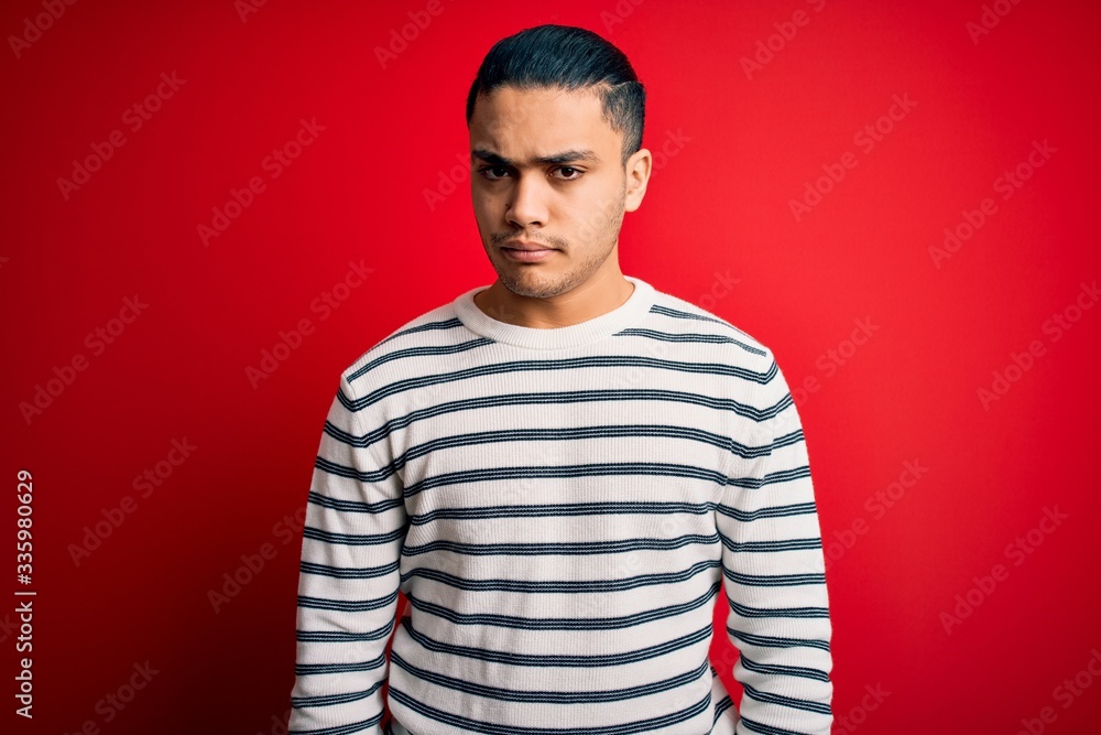 Young brazilian man wearing casual striped t-shirt standing over isolated red background skeptic and nervous, frowning upset because of problem. Negative person. Young brazilian man wearing casual striped t-shirt standing over isolated red background skeptic and nervous, frowning upset because of problem. Negative person.