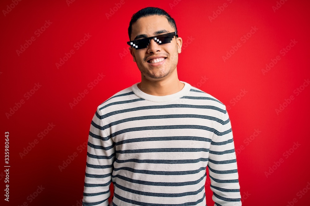 Young brazilian man wearing funny thug life sunglasses over isolated red background with a happy and cool smile on face. Lucky person. Young brazilian man wearing funny thug life sunglasses over isolated red background with a happy and cool smile on face. Lucky person.