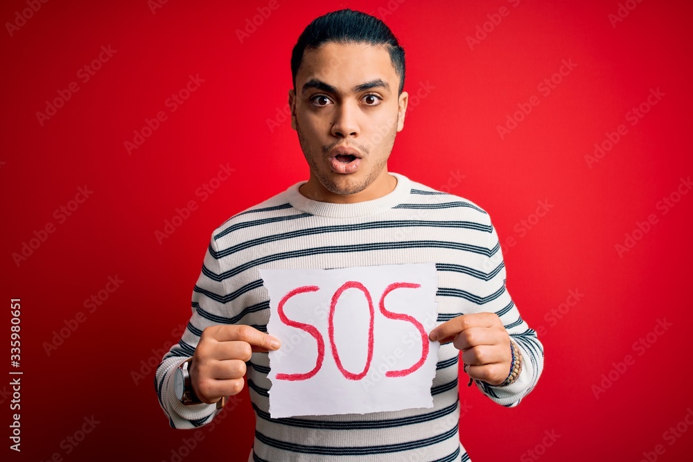 Young brazilian man with problem holding banner with sos message over red background scared in shock with a surprise face, afraid and excited with fear expression Young brazilian man with problem holding banner with sos message over red background scared in shock with a surprise face, afraid and excited with fear expression