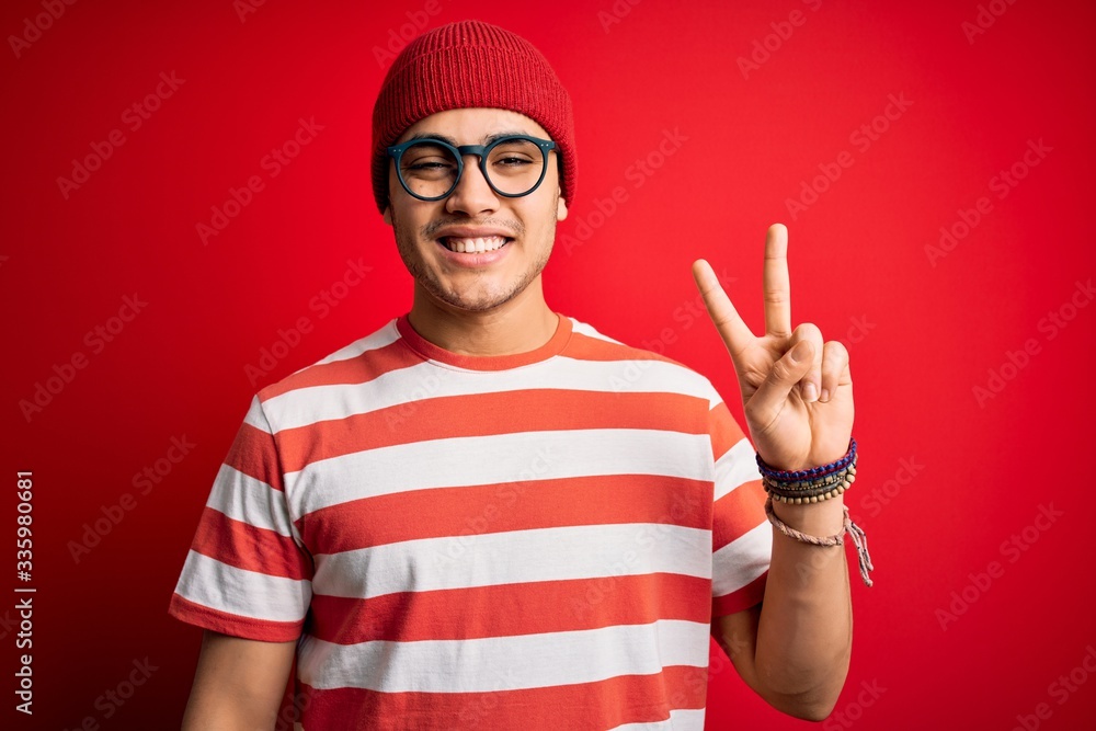 Naklejka premium Young brazilian man wearing casual striped t-shirt and glasses over isolated red background smiling with happy face winking at the camera doing victory sign. Number two.