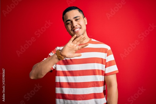 Young brazilian man wearing casual striped t-shirt standing over isolated red background showing and pointing up with fingers number five while smiling confident and happy.