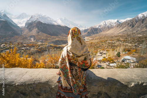 A woman wearing traditional dress sitting on wall and looking at Hunza valley in autumn season, Gilgit Baltistan in Pakistan