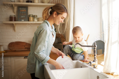 Indoor shot of cute positive young European female with ponytail posing in kitchen interior washing dishes, her adorable baby son sitting at sink, turning on tap, helping mother to do housework