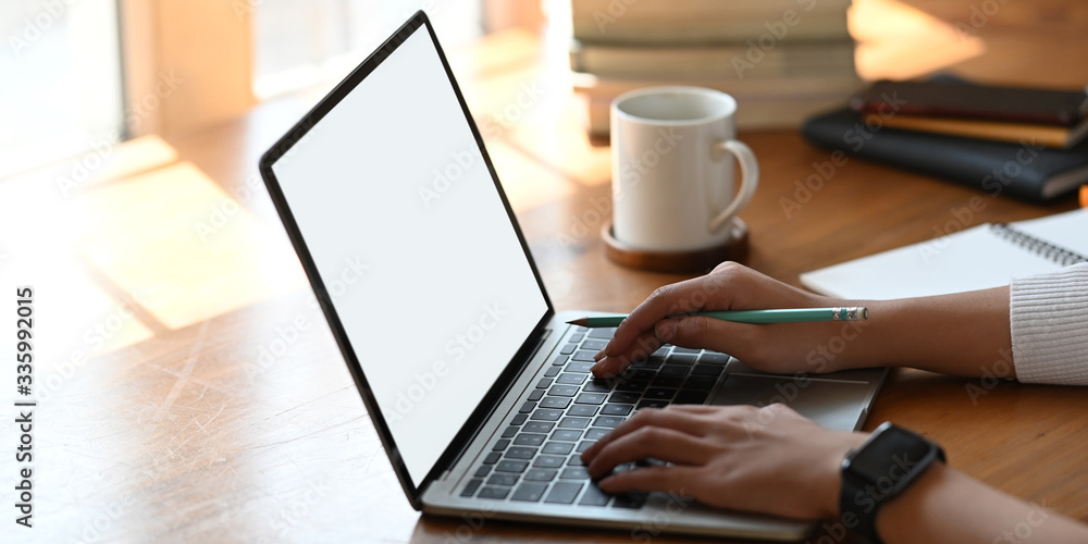 Cropped image of beautiful woman using a computer laptop with white blank screen while sitting at the white working desk over comfortable living room as background. Cropped image of beautiful woman using a computer laptop with white blank screen while sitting at the white working desk over comfortable living room as background.
