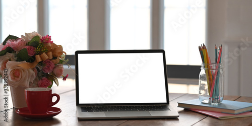 Laptop putting on working desk with bunch of flowers, notebook and pencil holder over comfortable living room as background.