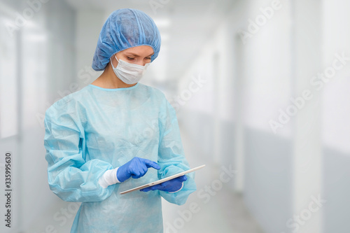 Female doctor in protective suit: medical mask, hat and robe. The woman uses a digital tablet and displays disease statistics on the screen.