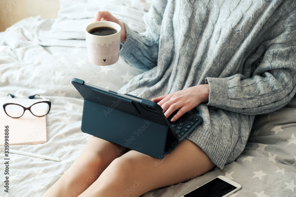 Obraz premium Young woman working on tablet and drinking coffee in bed at home. Cozy, hygge style. Online shopping, working home, freelance, online learning, studying concept. Distance education