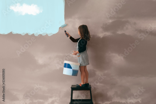little girl uses a can of paint to color the wall of the room from cloudy gray to clear blue sky - positive attitude, vibes and mentality concept photo
