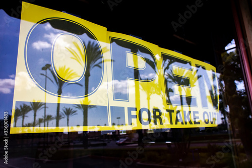 Close up of yellow sign with black letters in the window at restaurant stating they are Open For Take Out . The window reflects palm trees photo