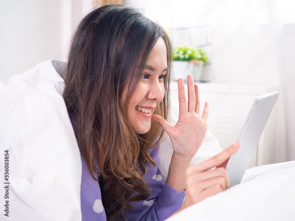 Bright-looking Asian woman is lying on the bed. Use the tablet to talk to video conferencing communicate and talk with people who are far away. social distance concept Bright-looking Asian woman is lying on the bed. Use the tablet to talk to video conferencing communicate and talk with people who are far away. social distance concept
