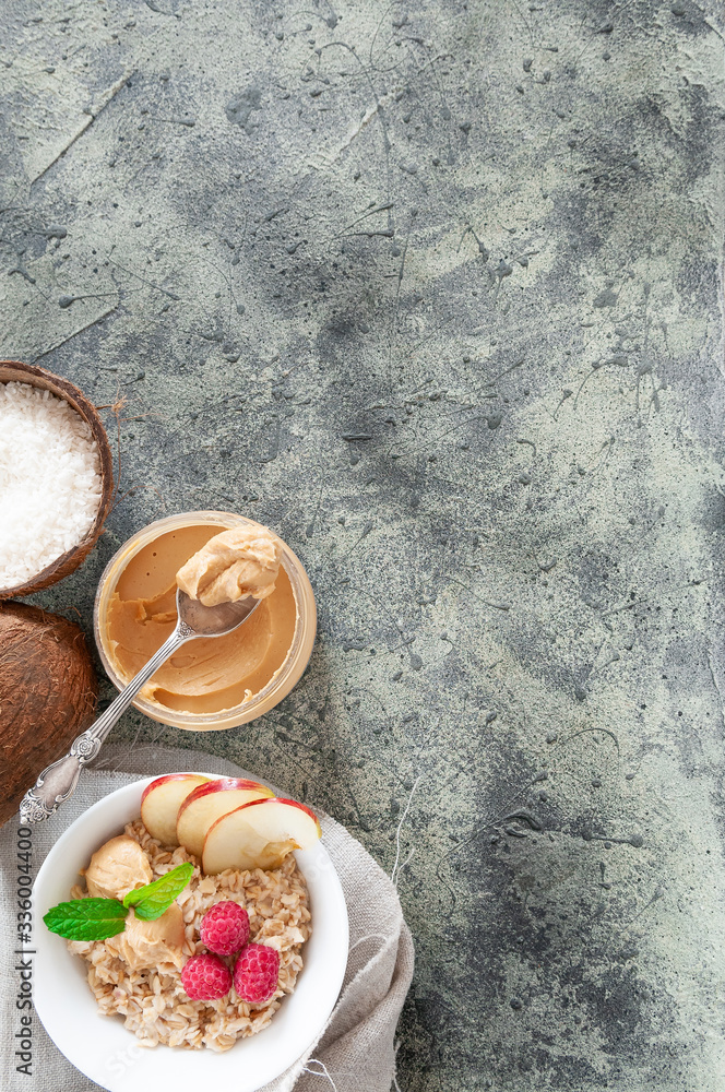 Oatmeal in a white plate with peanut butter, raspberries, apple slices and mint. Near a jar of peanut butter. Coconuts and a spoon. Place for text. Gray concrete background. Oatmeal in a white plate with peanut butter, raspberries, apple slices and mint. Near a jar of peanut butter. Coconuts and a spoon. Place for text. Gray concrete background.