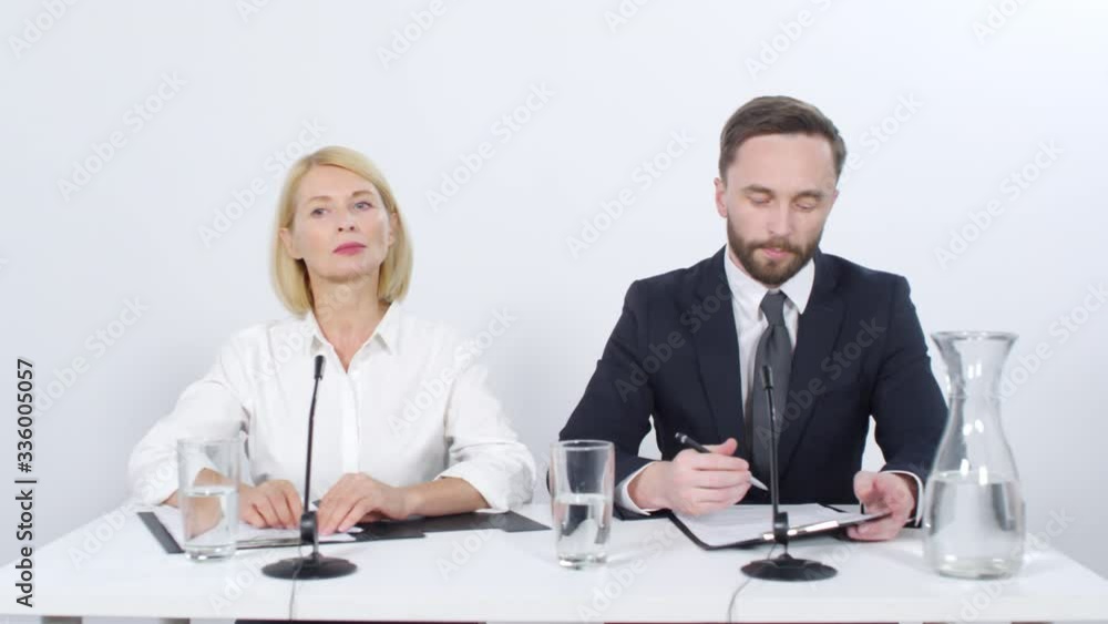 Young businessman giving a speech and drinking water during press conference while middle aged female colleague Young businessman giving a speech and drinking water during press conference while middle aged female colleague