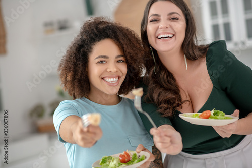 Two cheerful girlfriends offering to try vegetable salad.