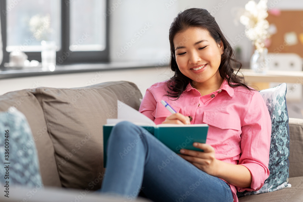 people and leisure concept - happy smiling asian young woman with diary or notebook sitting on sofa at home people and leisure concept - happy smiling asian young woman with diary or notebook sitting on sofa at home