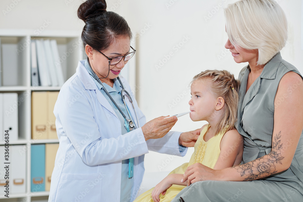 Pediatrician checking temperatire of little girl sitting on laps of her mother Pediatrician checking temperatire of little girl sitting on laps of her mother