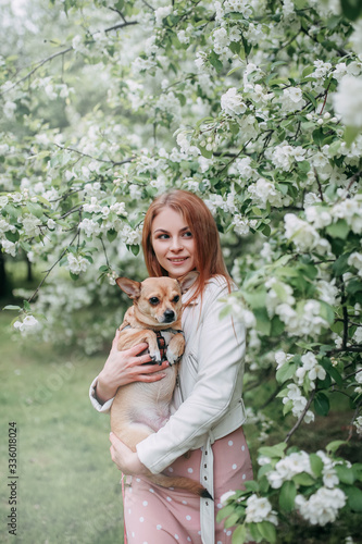 Beautiful gentle dreamy sweet pretty happy girl in a delicate dress and hat walking with her pet, with a dog in the park among the blooming apple trees in spring, friendship