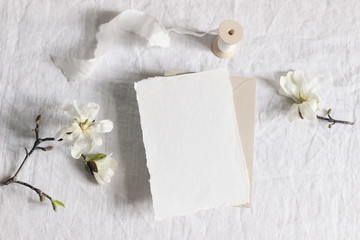 Wedding stationery mock-up scene. Blank vertical greeting card, envelope on linen tablecloth background with white magnolia stellata tree branches and ribbon. Feminine still life. Flat lay, top view.