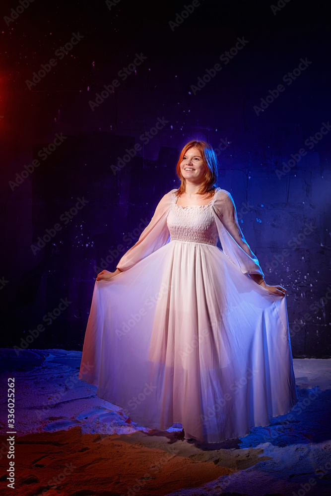 Pretty young sexy model female with blonde hair in amazing long white dress and posing in dark studio