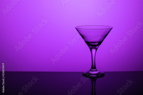 Glass goblet without wine on a thin leg stands on a mirror surface. Back light. Purple background. Copy space.
