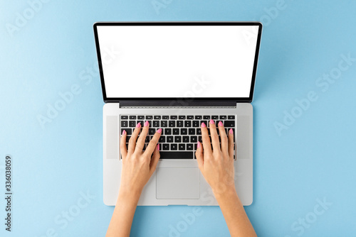 Woman’s hands working on laptop with empty screen on blue background. Office desktop. Top view