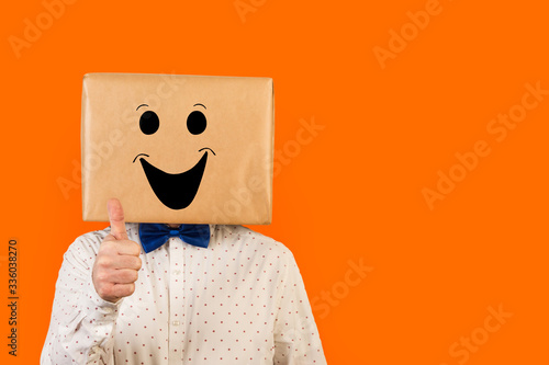 Hombre con caja de cartón en la cabeza con gesto positivo y feliz y el dedo pulgar de la mano hacia arriba OK sobre fondo naranja liso y aislado. Vista de frente y de cerca. Copy space